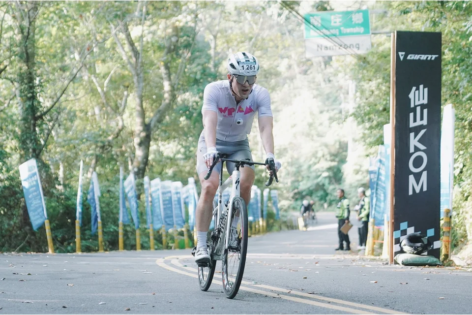 白衣選手騎近仙山 KOM 立牌，兩側插滿藍色活動旗幟