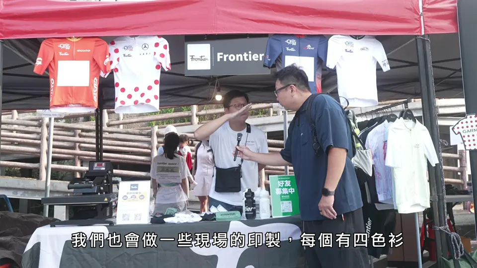 主持人在掛滿車衣的攤位前與廠商交談。