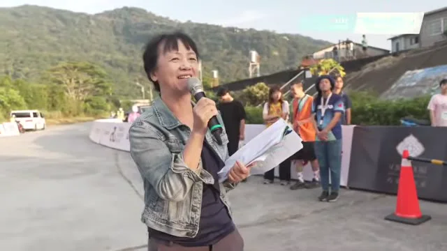 主持人於戶外會場持麥克風與資料發言，背景有工作人員。