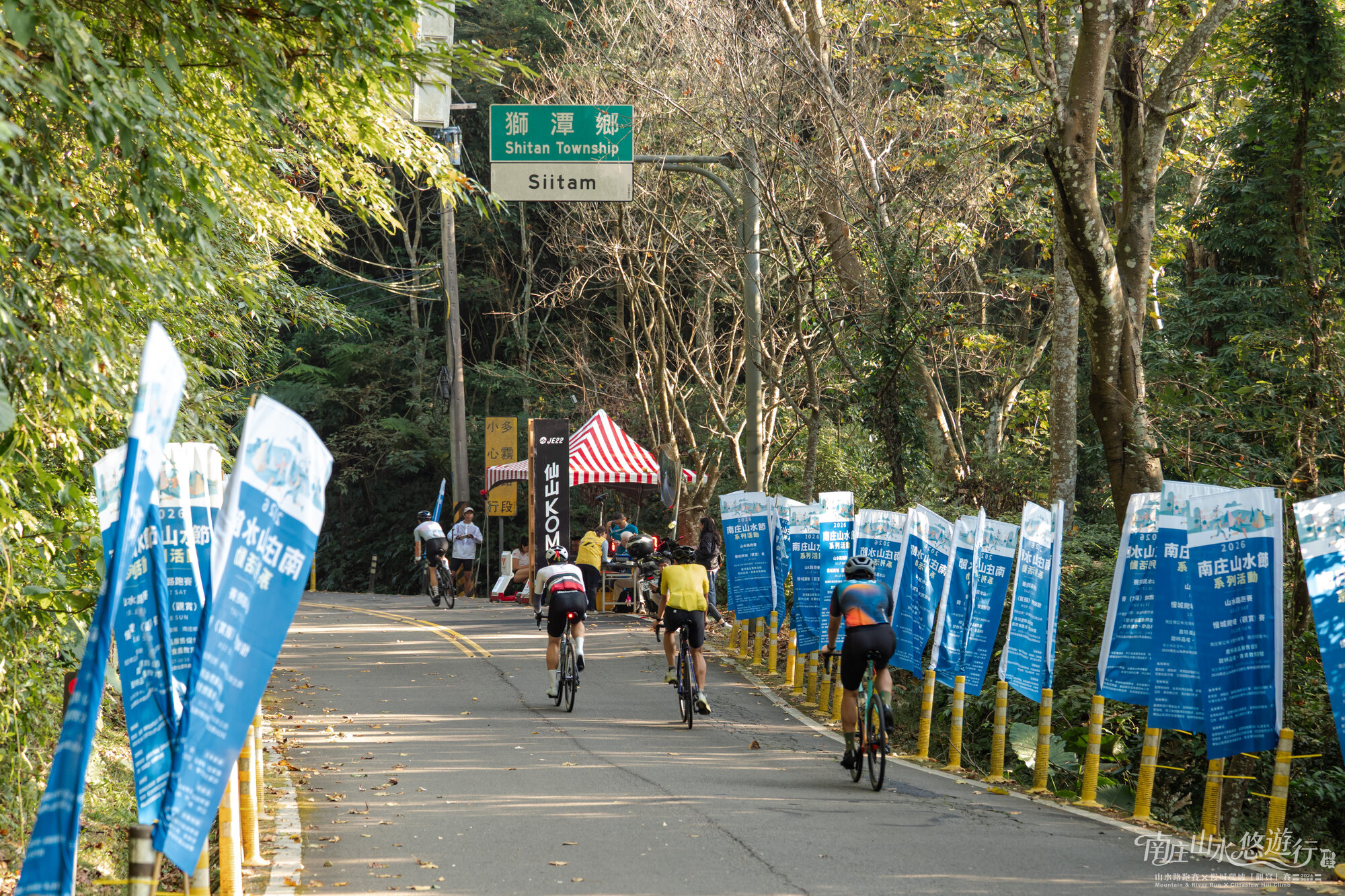 自行車選手沿藍色活動旗幟排列的山路騎向 KOM 路段的實景照片